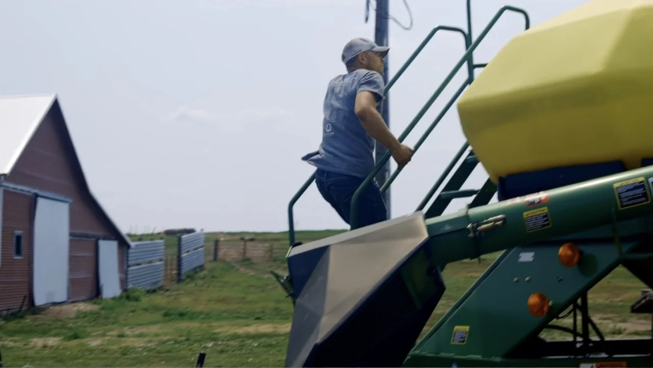 Common Ground (2000) movie still: medium — A rural agricultural setting with a large green tractor and a red metal building. A man in a blue t-shirt…