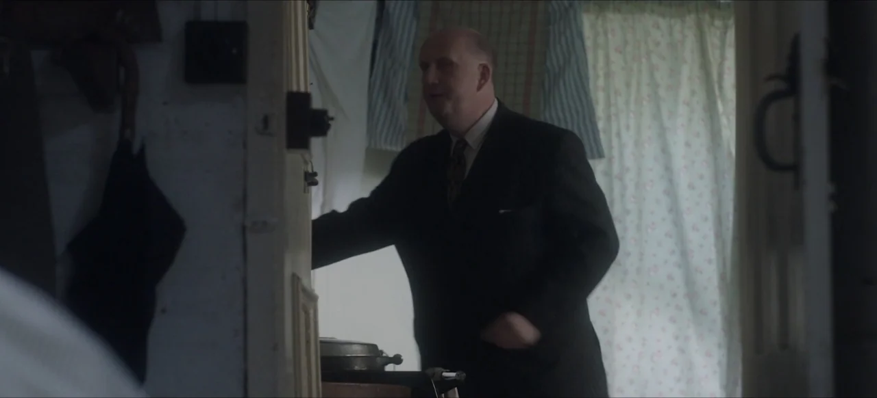 We Go In at Dawn (2020) movie still: medium — A dimly lit domestic hallway with white walls and floral curtains in the background. A middle-aged man…