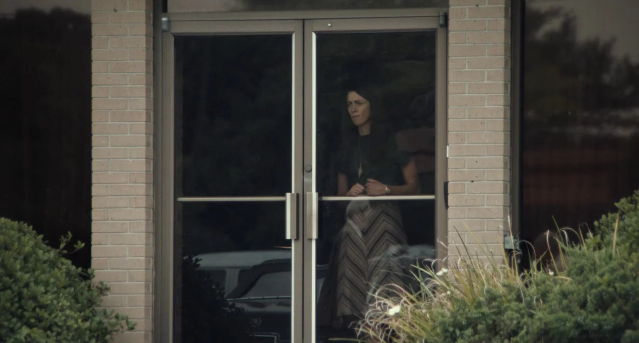 Christine (2016) movie still: medium — A brick-fronted building entrance features double glass doors reflecting a parked car. A woman stands inside…