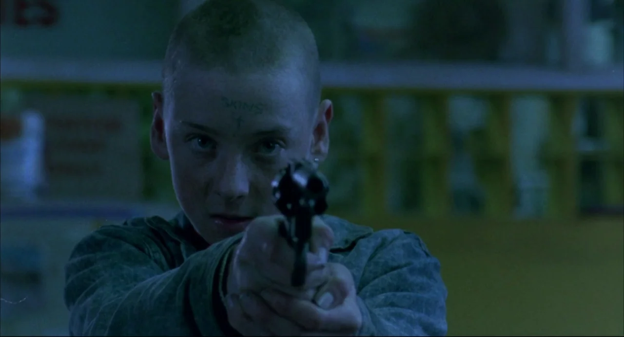 Romper Stomper (1992) movie still: medium — A dimly lit indoor retail space with blurred shelves in the background. A young person with a shaved…