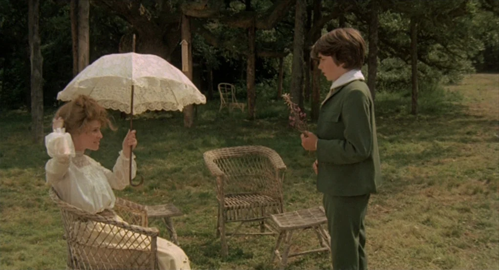 The Go-Between (1971) movie still: medium — A grassy clearing surrounded by trees contains antique wicker furniture in a secluded natural setting. A…