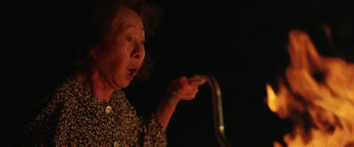 Minari (2020) movie still: medium — An elderly woman sits outside at night near an open fire. She looks toward the flames with her mouth slightly…