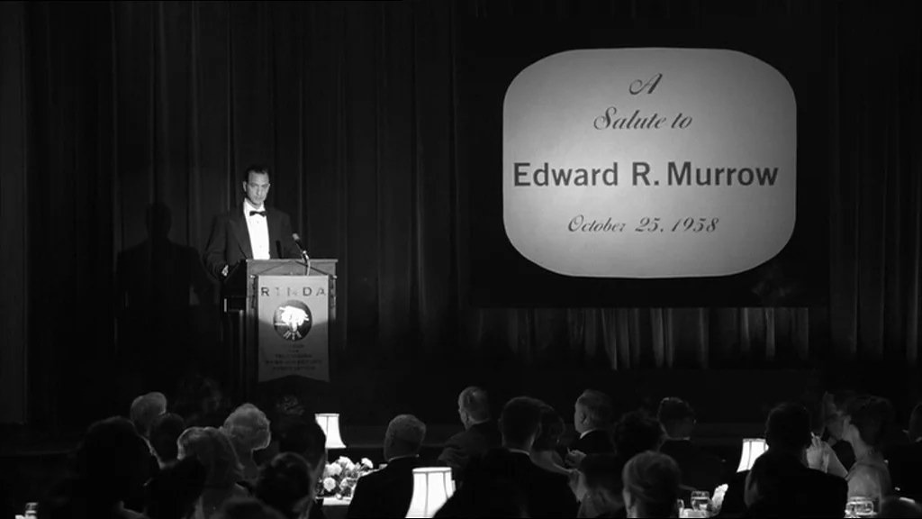 Good Night And Good Luck (2005) movie still: wide — A formal event room with dark curtains behind a wooden lectern on a stage. A man in a tuxedo…