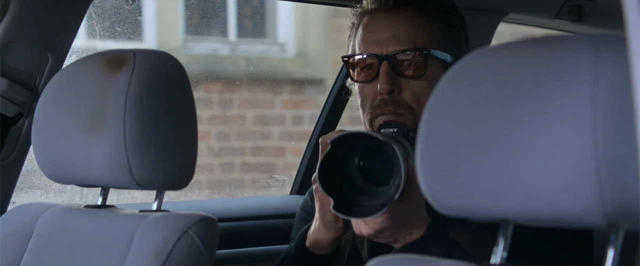 The Gentlemen (2020) movie still: medium — A man sits inside a vehicle while looking through a large camera lens. He wears glasses and holds the…