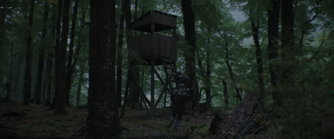 Wolf Man (2025) movie still: wide — A dense, dark woodland filled with tall, moss-covered trees and thick undergrowth. Two individuals with large…