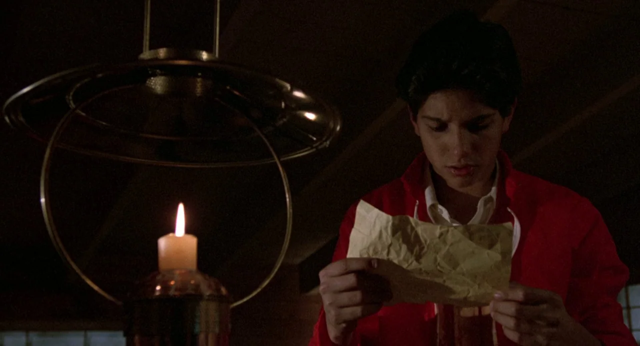 The Karate Kid (1984) movie still: medium — A dimly lit indoor space is illuminated by a single flickering candle. A young man in a red jacket reads…