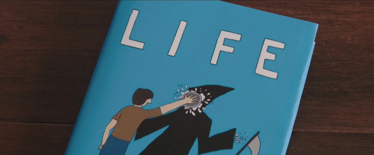 Dean (2016) movie still: closeup — A book cover with a bright blue background sits on a dark wooden surface. The cover features a cartoon graphic of…