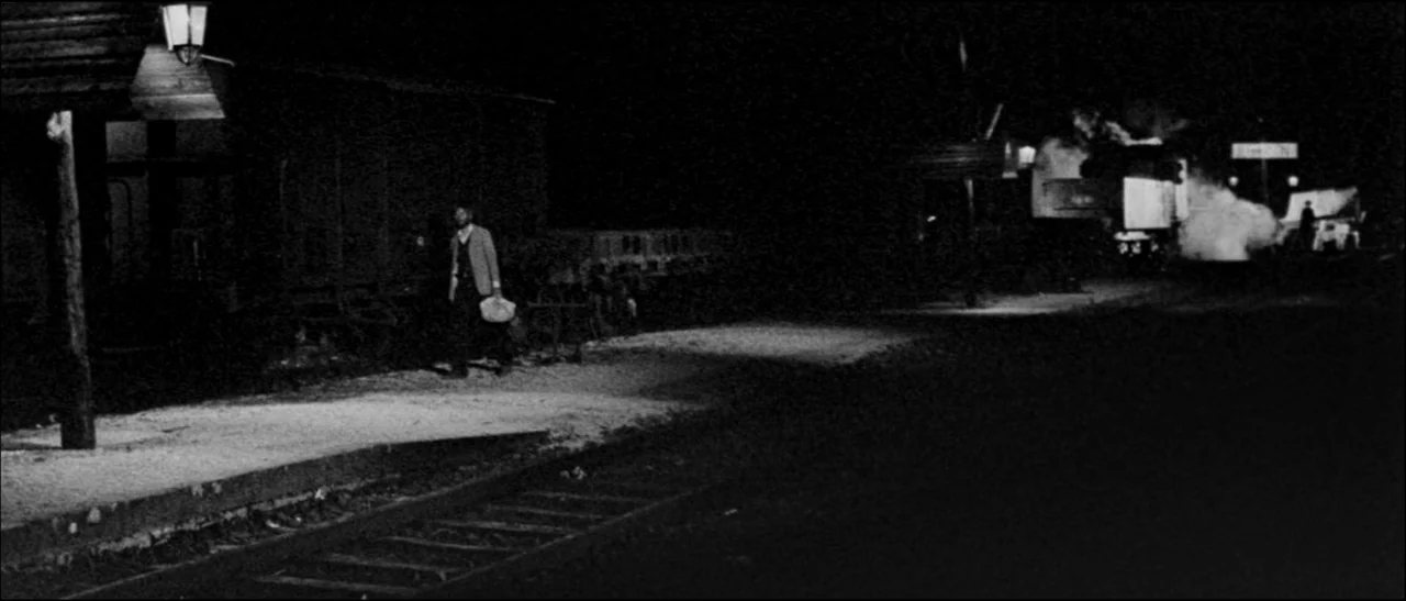 The Grand Duel (1972) movie still: wide — A rural train station platform at night is lit by a single overhead lantern. A man carrying a small bag…
