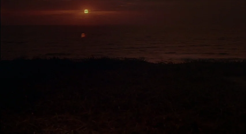 Mishima: A Life in Four Chapters (1985) movie still: establishing — A dark, silhouetted beach landscape sits before a calm ocean at sunset. The…