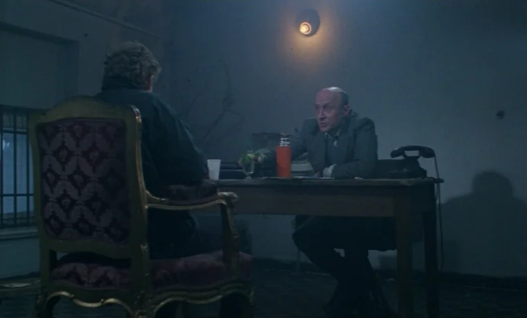 Ga-ga: Glory to the Heroes (1986) movie still: medium — A dim room with a lone practical light fixture mounted on the wall. Two men sit at a wooden…