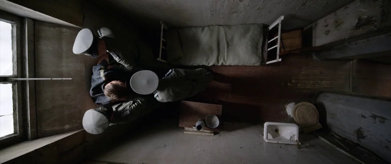 A Hidden Life (2019) movie still: fullbody — A small stone room contains a metal cot, a low toilet, and a sink. Three uniformed men stand in a group…
