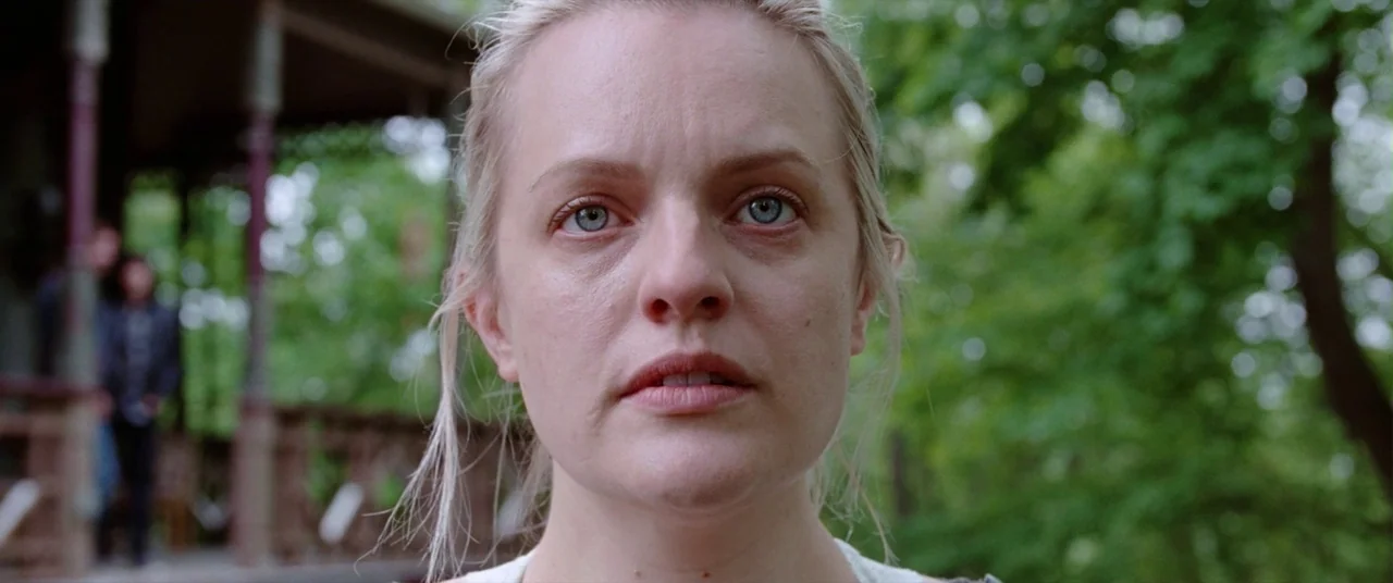 Her Smell (2018) movie still: closeup — A woman stands in front of a blurred porch structure surrounded by green trees. She looks forward directly…