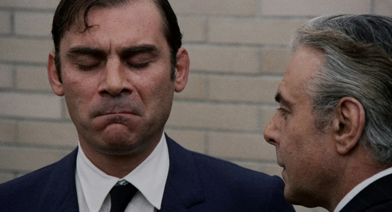 Investigation of a Citizen Above Suspicion (1970) movie still: medium — Two men stand in front of a neutral-toned brick wall during the daytime. The…