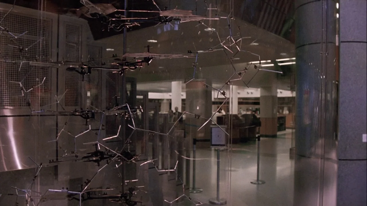 The Manchurian Candidate (2004) movie still: wide — A glass display containing a complex metal molecular model stands in a wide lobby. Through the…