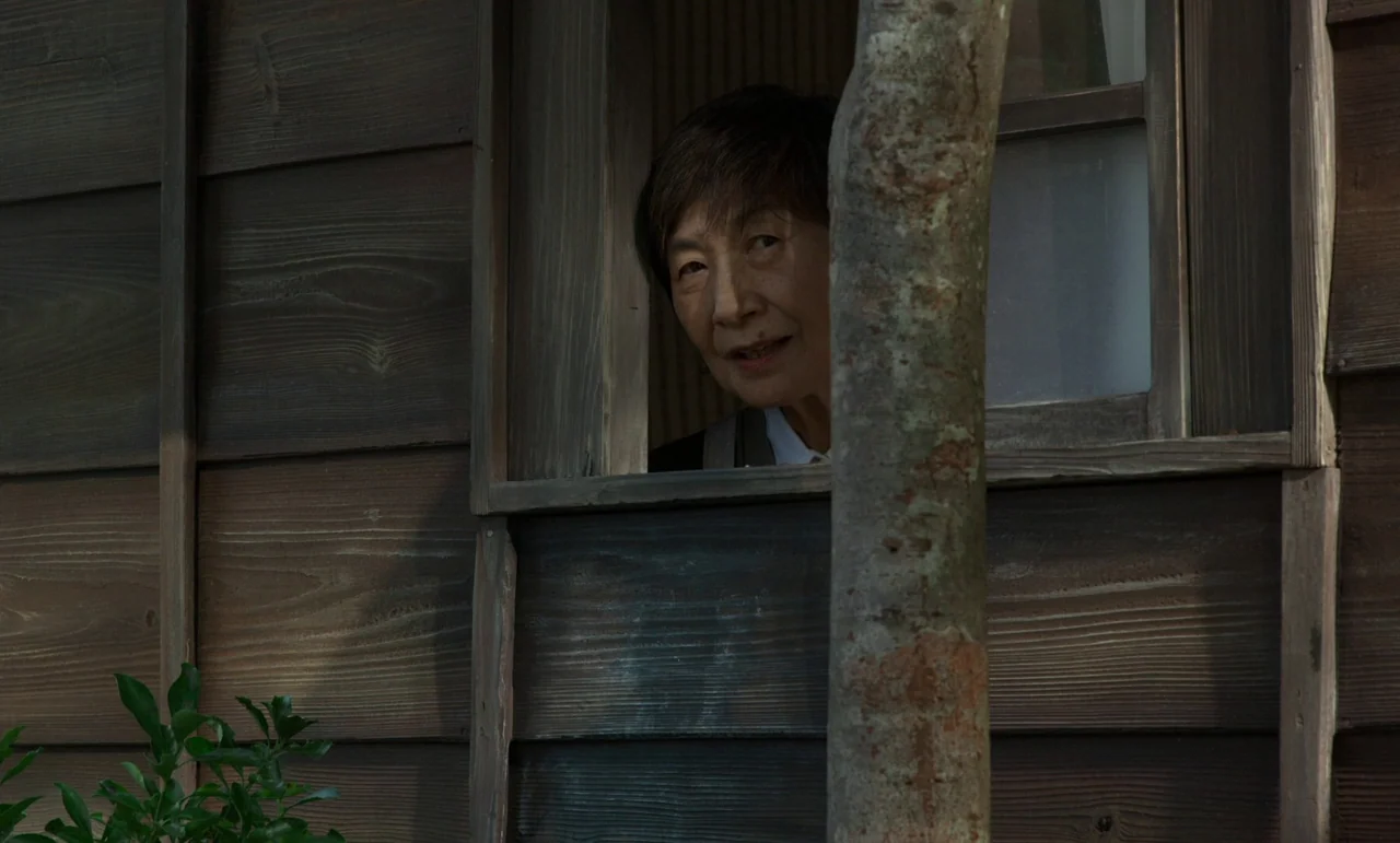 Like Someone In Love (2012) movie still: medium — A wooden wall with horizontal dark cedar planks frames a rectangular window opening. An elderly…