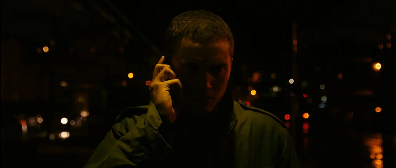 Love (2015) movie still: medium — A dark outdoor urban setting at night features distant bokeh streetlights. A man in an olive green jacket holds a…