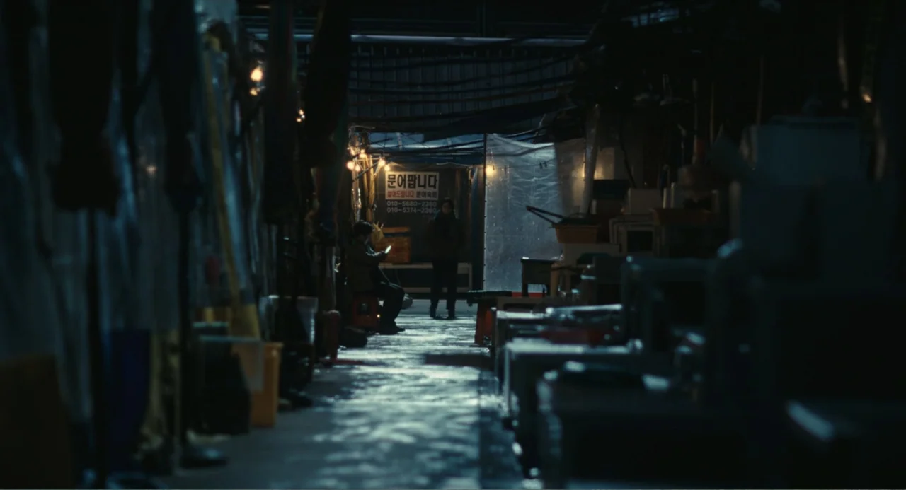 Winter in Sokcho (2025) movie still: wide — A narrow, cluttered outdoor market aisle at night features damp surfaces and plastic sheeting. A person…