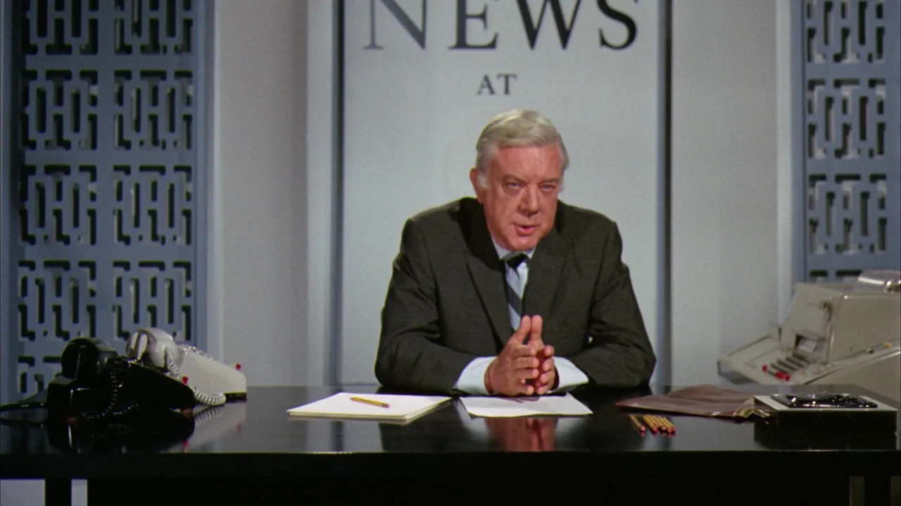 Willy Wonka & the Chocolate Factory (1971) movie still: medium — A television news desk set features a backlit sign reading 'NEWS AT'. A man in a…