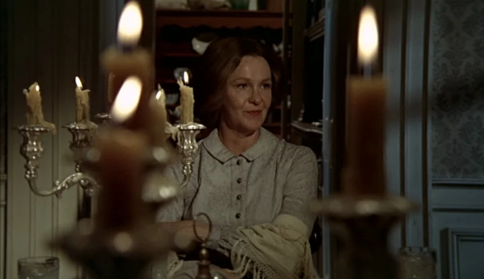 The Beguiled (1971) movie still: medium — A woman sits at a wooden table in a dimly lit domestic dining space. She looks ahead while blurred candles…