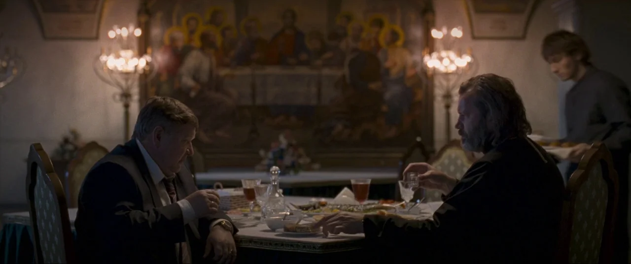 Leviathan (2014) movie still: medium — A dimly lit room contains a table set for dinner beneath a large religious painting. Two men face each other…