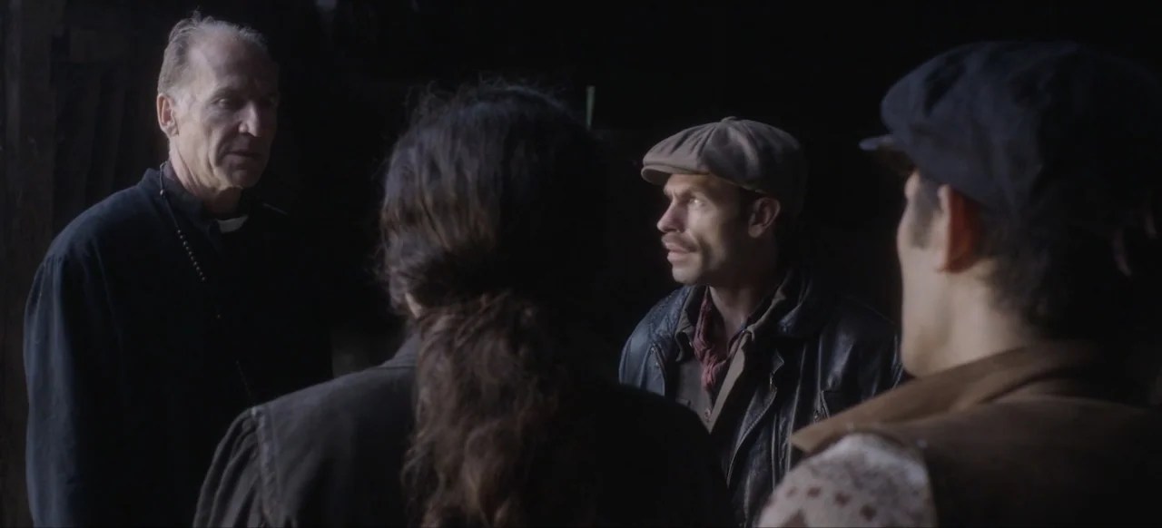 We Go In at Dawn (2020) movie still: medium — A dimly lit barn interior features three men in 1930s attire standing close together. An older man in…