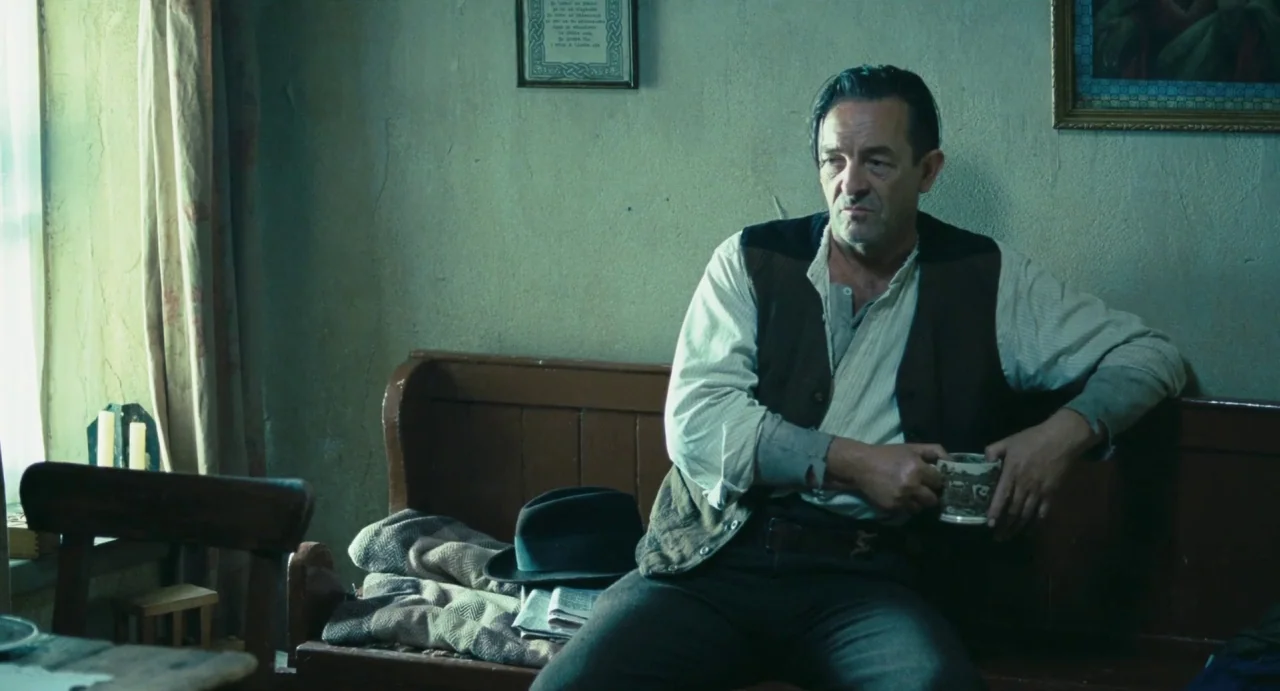 Jimmy’s Hall (2014) movie still: medium — A rustic interior features a plain plaster wall decorated with framed religious prints. A middle-aged man…