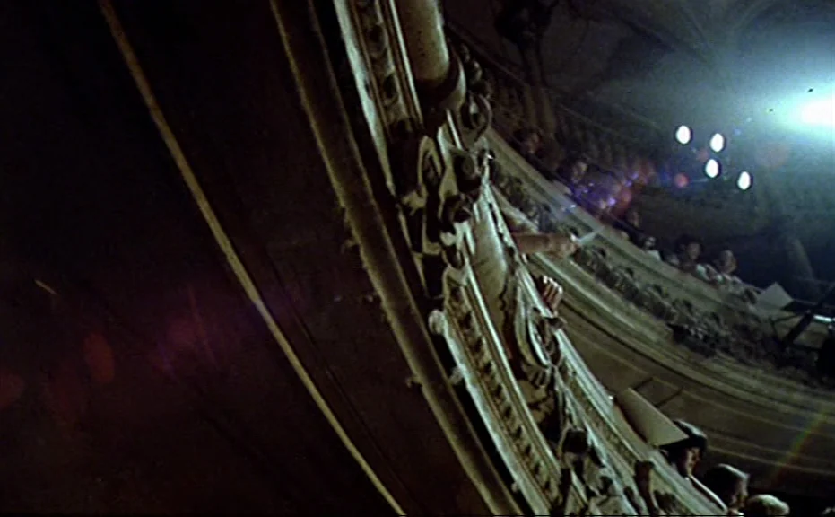 Diva (1981) movie still: wide — A dark, ornate theater balcony with intricate wood carvings dominates the foreground. Silhouetted figures sit in the…