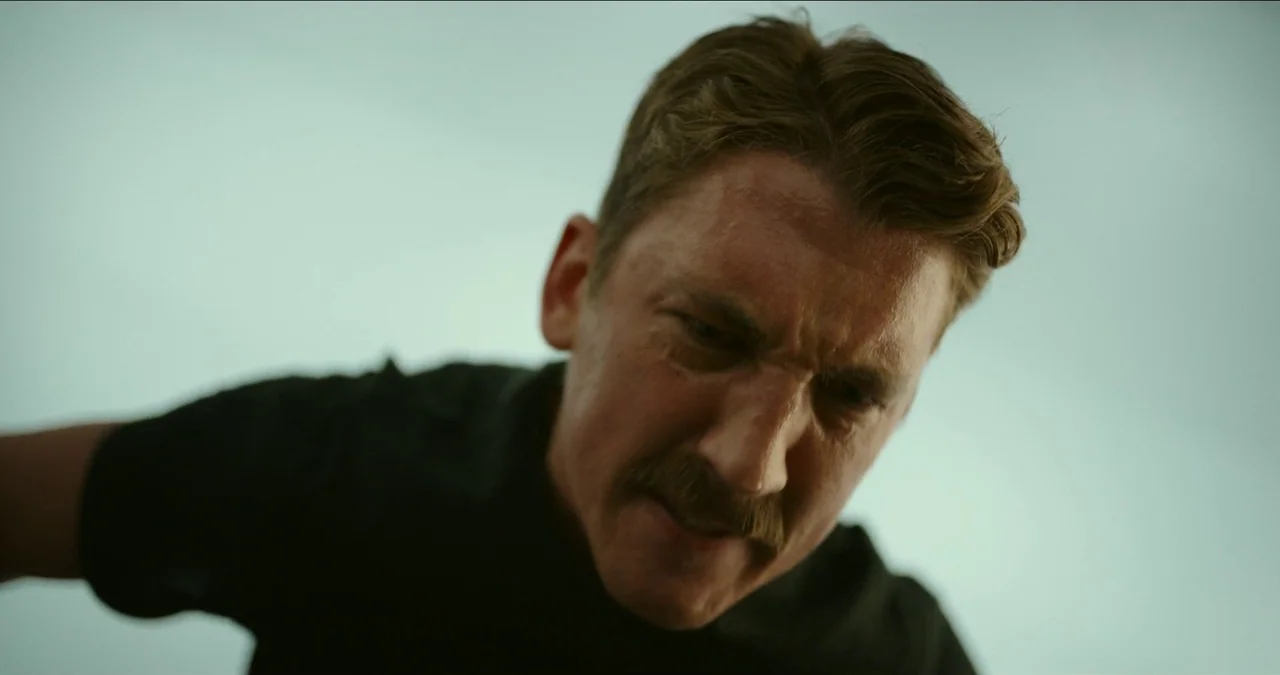 Top Gun: Maverick (2022) movie still: closeup — A man looks directly downward against a bright, hazy, light blue sky background. The frame focuses…