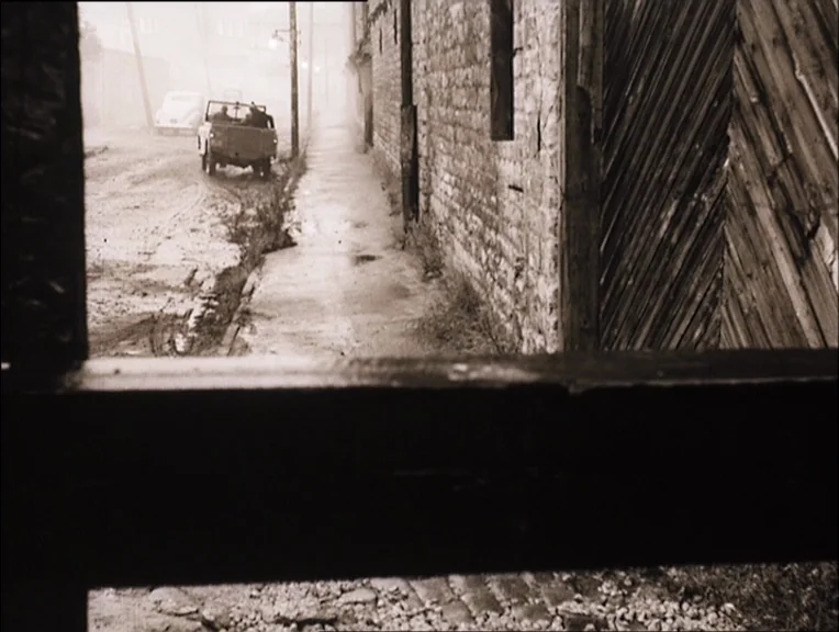 Stalker (1979) movie still: wide — A wet cobblestone road stretches past a weathered brick wall and wooden structure. A vintage automobile drives…