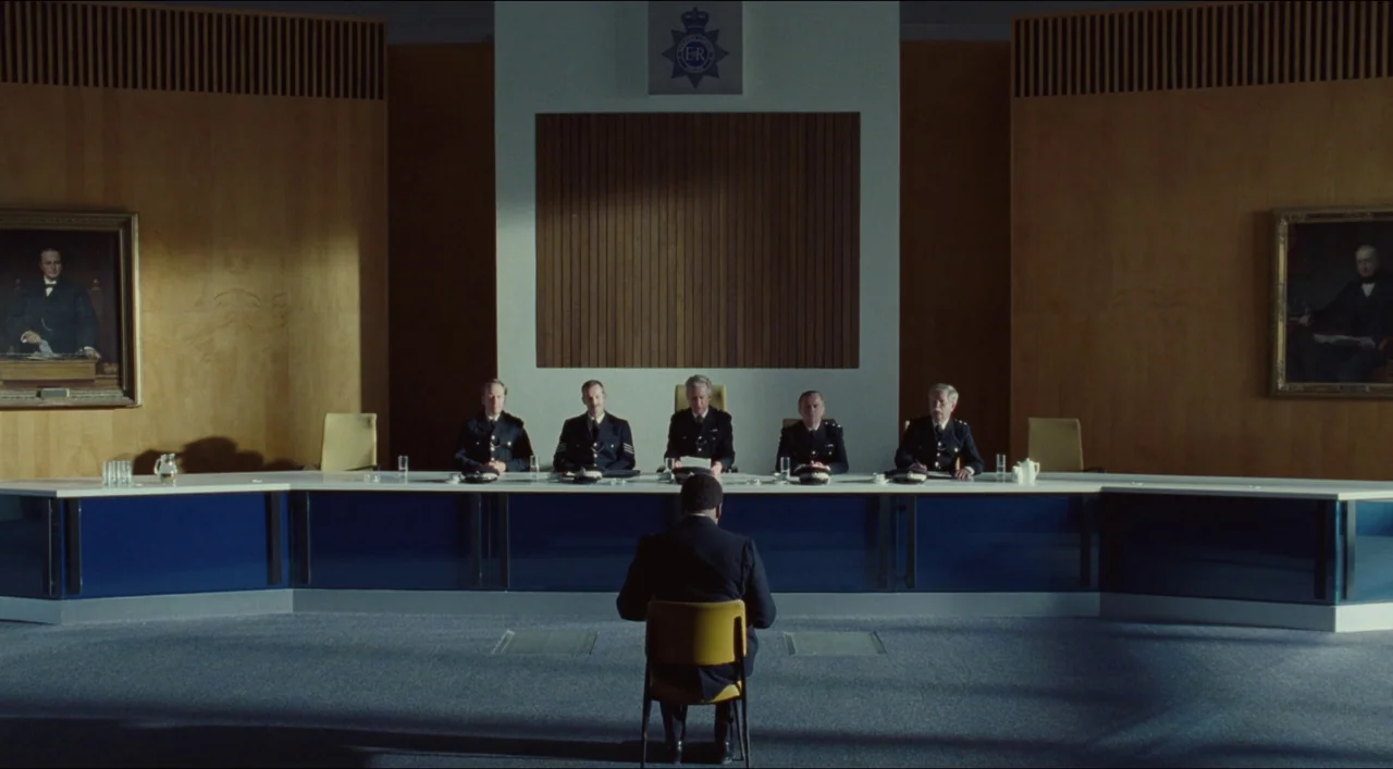 Red, White & Blue (2020) movie still: wide — A modern hearing room features a long white desk and wood-paneled walls. One man sits alone in a yellow…