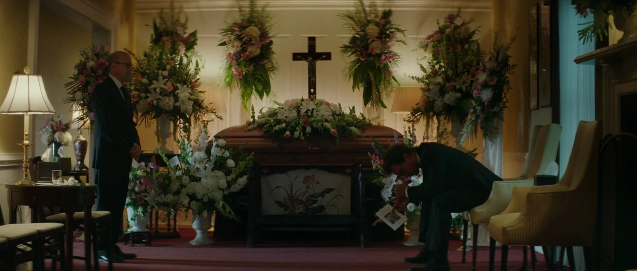 The Sea of Trees (2015) movie still: wide — A funeral parlor contains a wooden casket centered beneath a crucifix on the wall. A man stands silently…