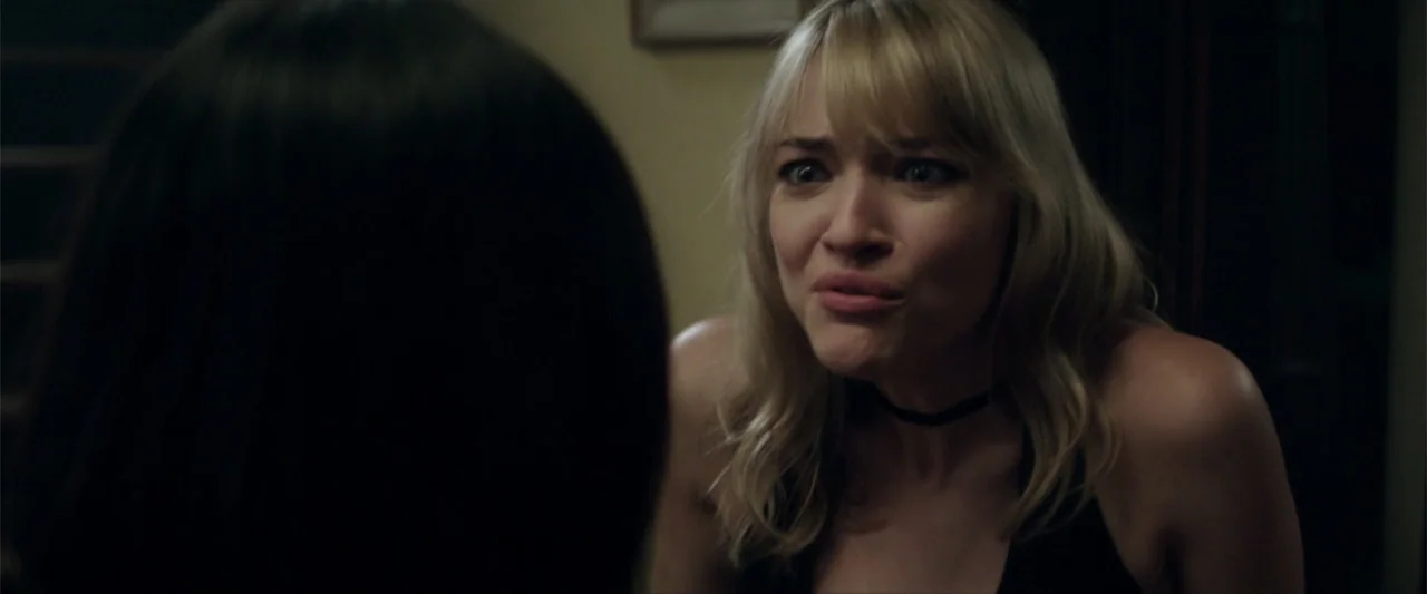 Truth or Dare (2018) movie still: medium — A dimly lit indoor space with plain wall surfaces in the background. A blonde woman with an expressive…