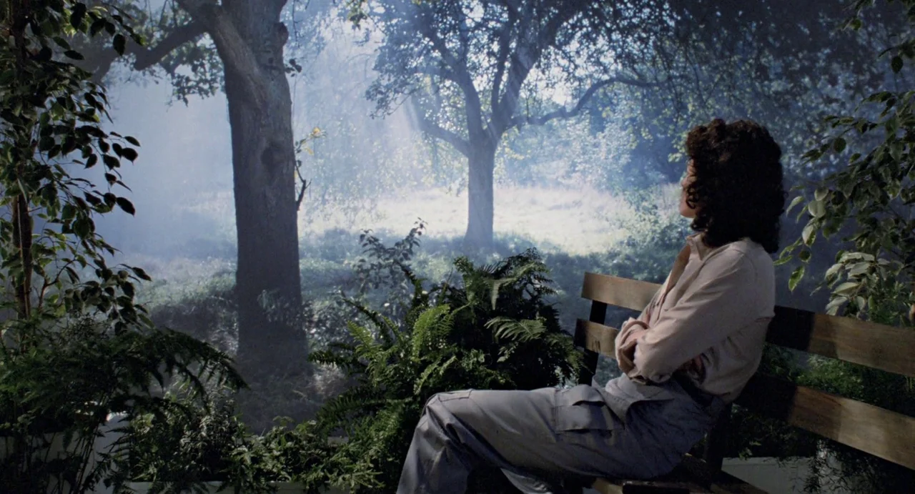 Aliens (1986) movie still: medium — A woman sits on a wooden bench surrounded by dense foliage and ferns. Thick trees fill the background as light…