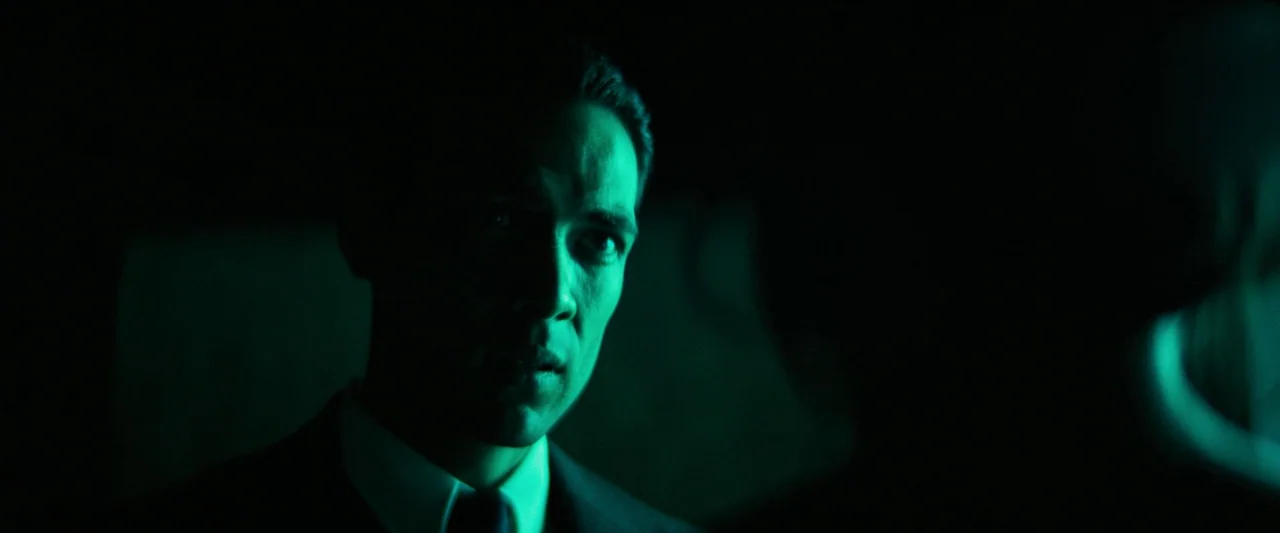 Babylon (2022) movie still: closeup — A man stands in a dark environment with very limited lighting. Only half of his face is visible under a strong…