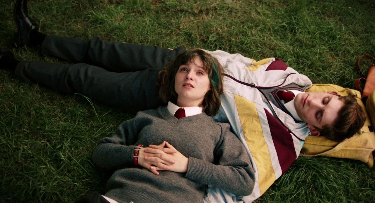 Dating Amber movie still: medium — Two teenagers in school uniforms lie flat on a patch of lush green grass. They are positioned side by side with…
