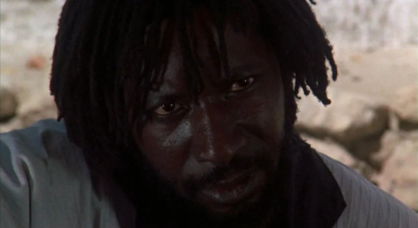 The Passenger (1975) movie still: closeup — A man stands outdoors in a bright, arid environment with rocky textures visible. The man stares directly…