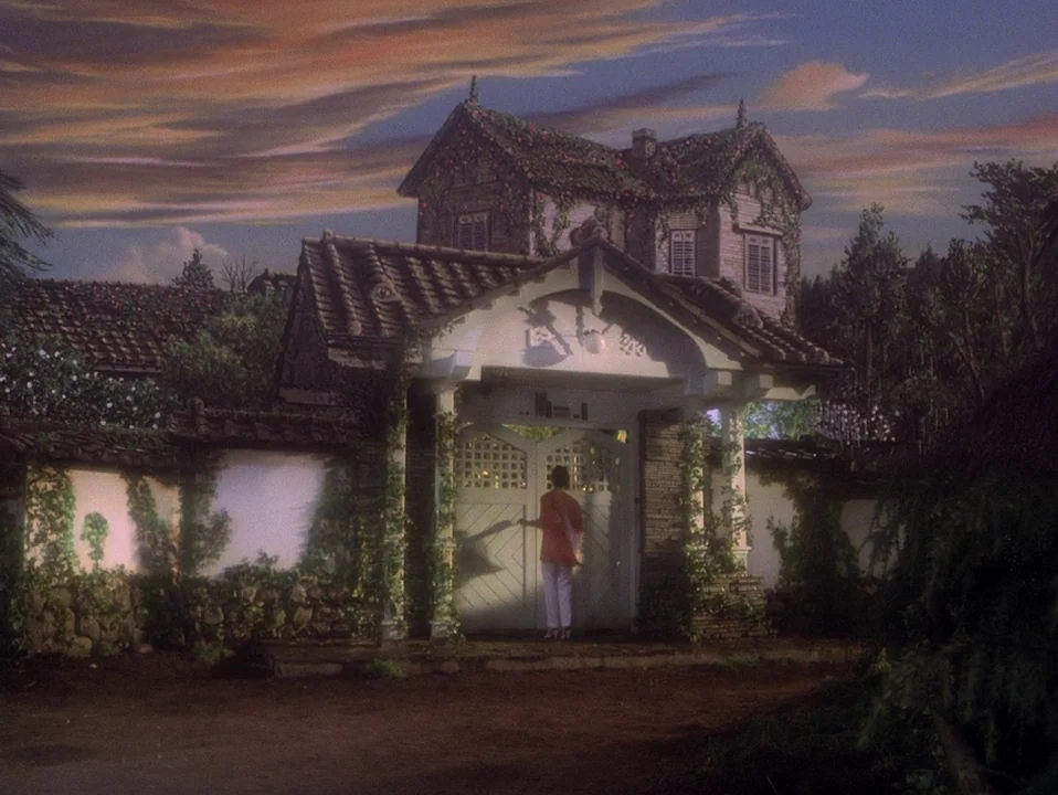 House (Hausu) (1977) movie still: wide — A two-story villa sits behind a high plastered wall covered in climbing vines. A person in light clothing…