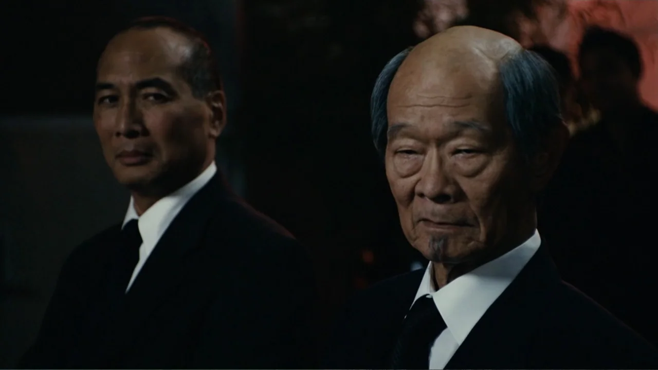 Click (2006) movie still: medium — A dark, formal interior space serves as the backdrop for two men in suits. The men stand side by side while…