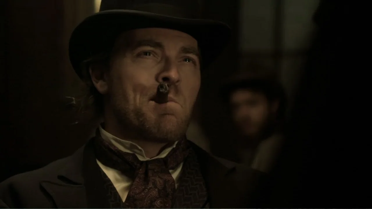 Dawn Rider (2012) movie still: closeup — A dimly lit 19th-century interior with a man visible in the foreground. The man wears period clothing and…