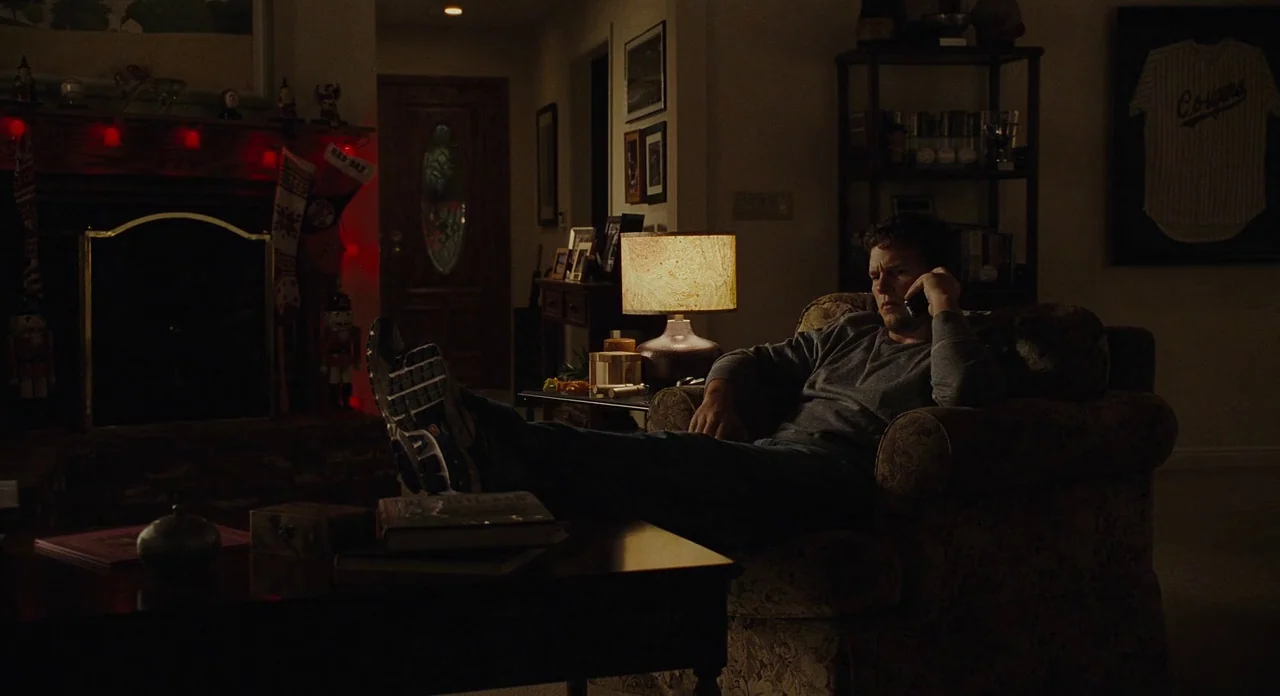 Moneyball (2011) movie still: medium — A dimly lit room with a fireplace containing red holiday lights and a framed jersey on the wall. A man sits…