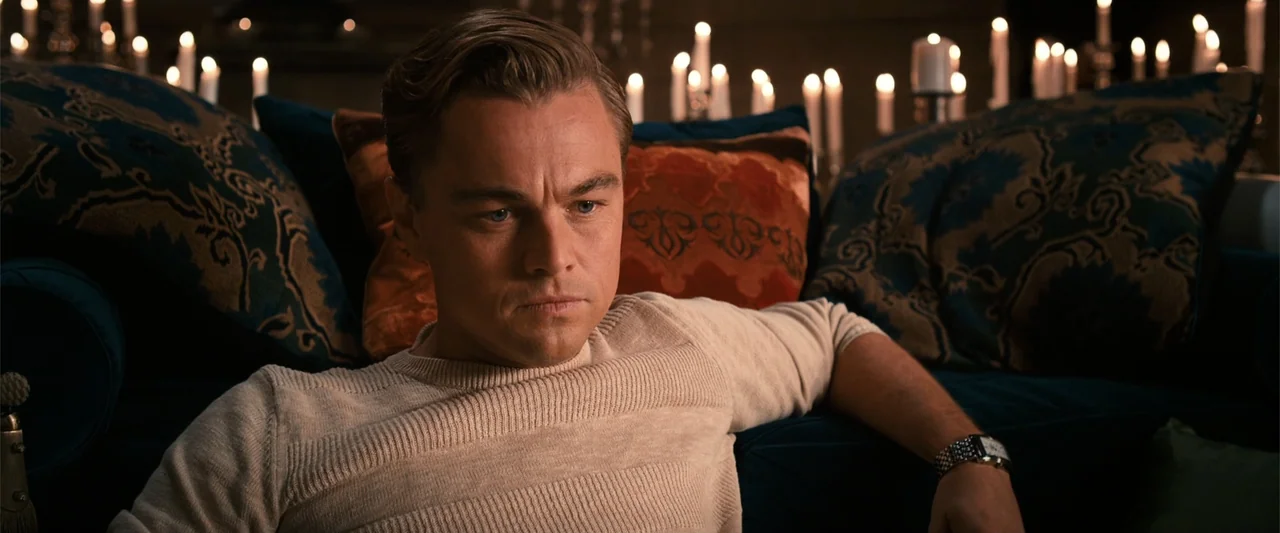 The Great Gatsby (2013) movie still: medium — A dimly lit room features a dark teal sofa adorned with orange and blue cushions. A man wearing a…