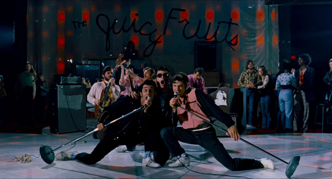 Phantom of the Paradise (1974) movie still: medium — A 1970s band performs on a white floor with a black-lettered backdrop. Members of the band sing…