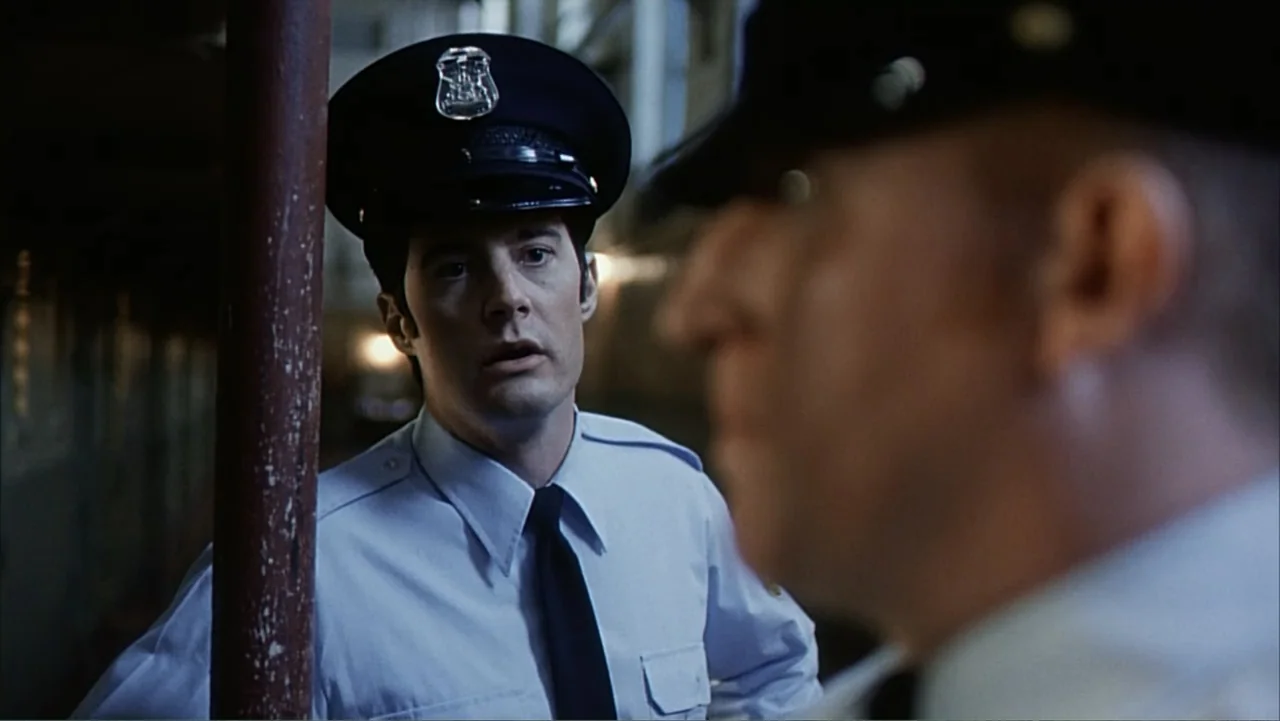Against the Wall (1994) movie still: medium — A dimly lit interior hallway with a vertical metal support pole in the foreground. A police officer in…