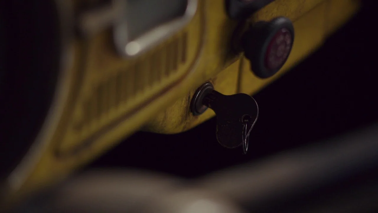 Bumblebee (2018) movie still: closeup — A metal dashboard of an older vehicle has a yellow paint finish with visible wear. A single ignition key is…