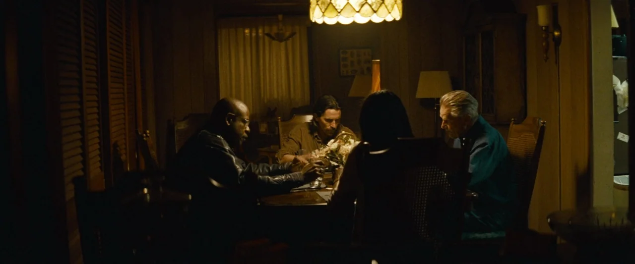 Out of the Furnace (2013) movie still: medium — A dark, wood-paneled room is lit only by a central pendant fixture over dinner. Four adults sit…