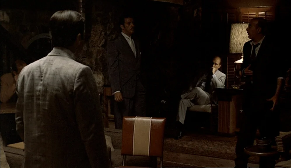 The Godfather Part II (1974) movie still: medium — A wood-paneled room is dimly lit by a single table lamp. Four men in formal business attire stand…