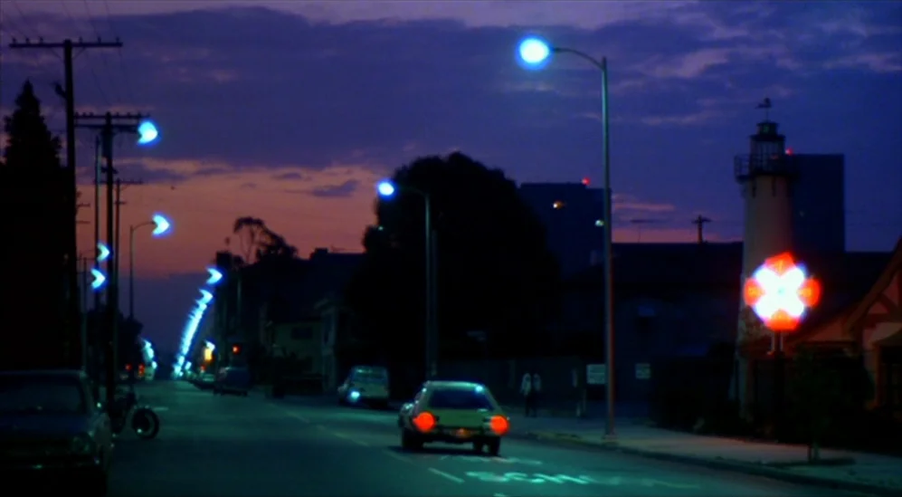American Gigolo (1980) movie still: establishing — A quiet suburban street at dusk with silhouettes of buildings and a lone lighthouse. A car…