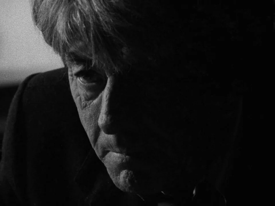 Following (1998) movie still: closeup — A close-up captures an elderly man in a dimly lit interior setting. He gazes downward with part of his face…