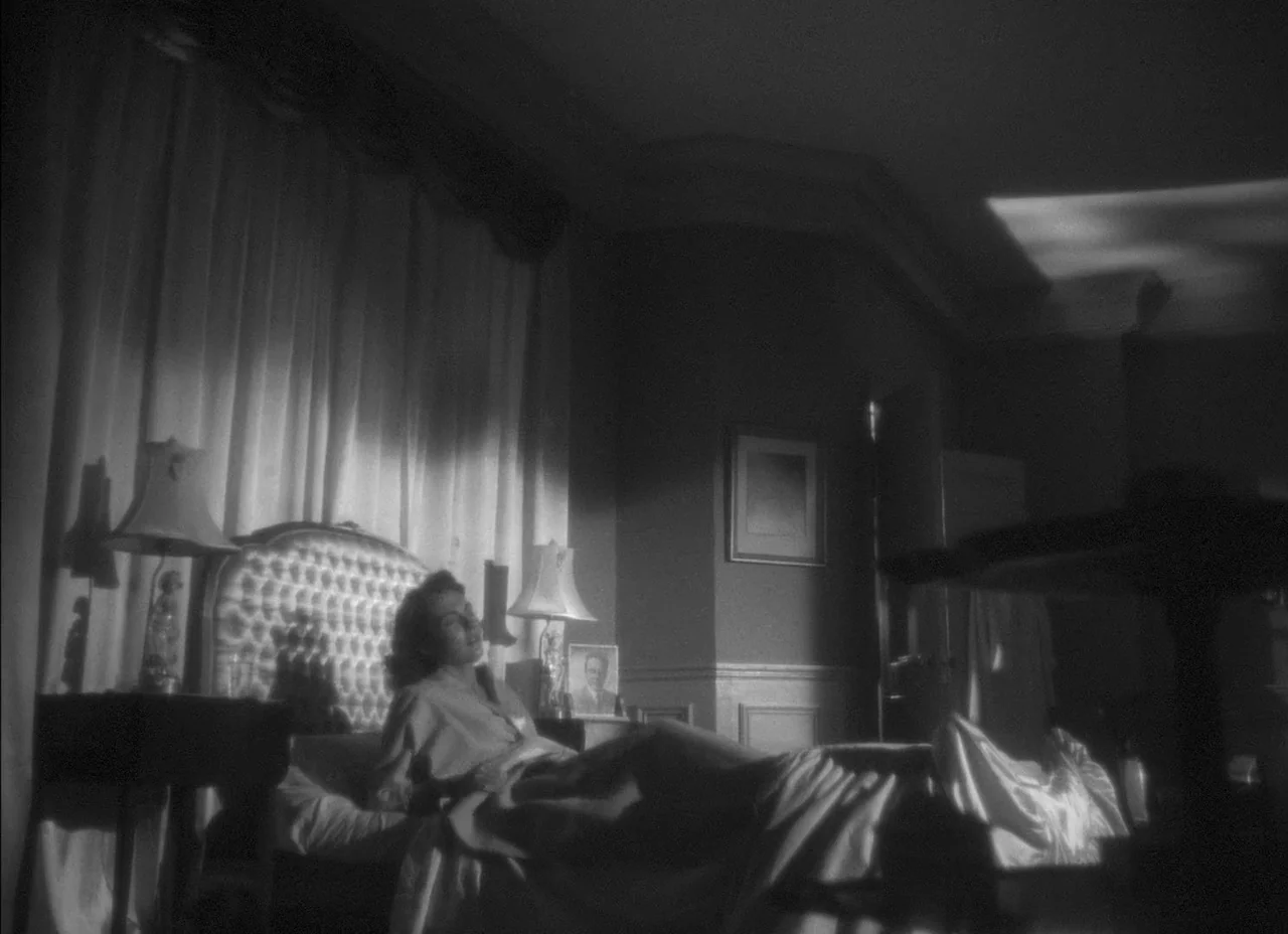 The Amazing Mr. X (1948) movie still: wide — A 1940s style room features a tufted headboard and heavy curtains. A woman rests in bed while…