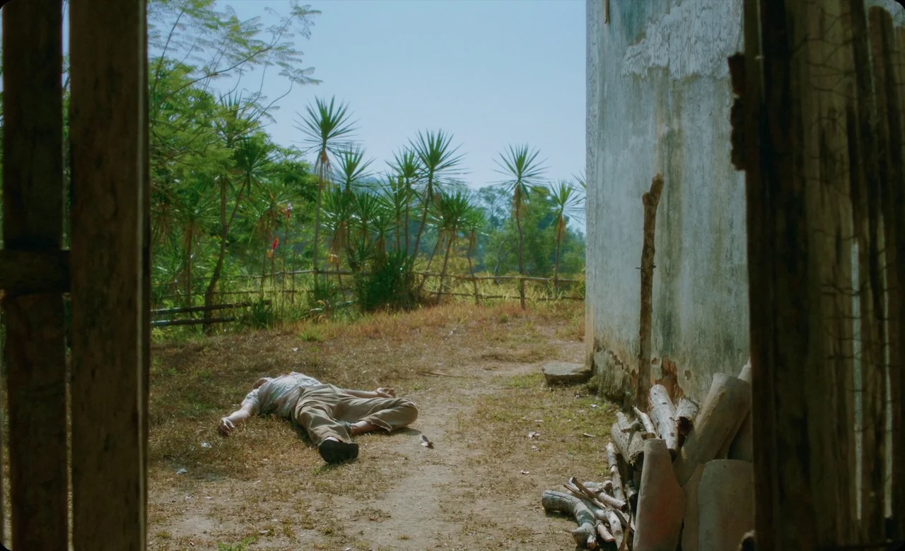 Eureka (2024) movie still: wide — An outdoor area with dry, sparse grass borders a weathered, pale gray concrete wall. A man in light clothing lies…
